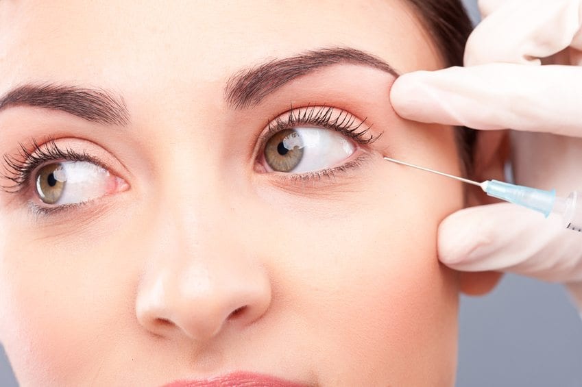 Close up of female face getting botox injection. Young woman is looking aside with concentration. Hand of doctor is holding a syringe near her eye