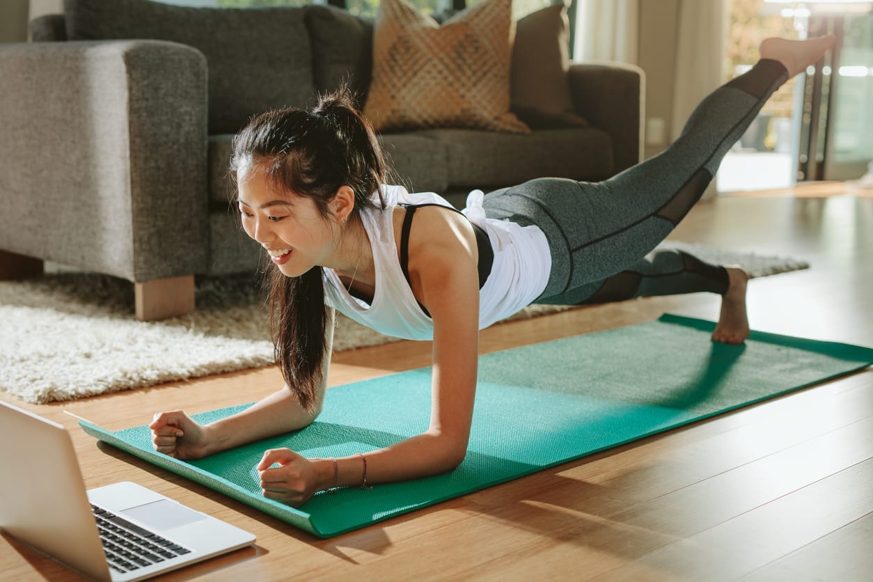 Smiling woman exercising at home and watching training videos on laptop. Chinese female doing planks with a leg outstretched and looking at laptop