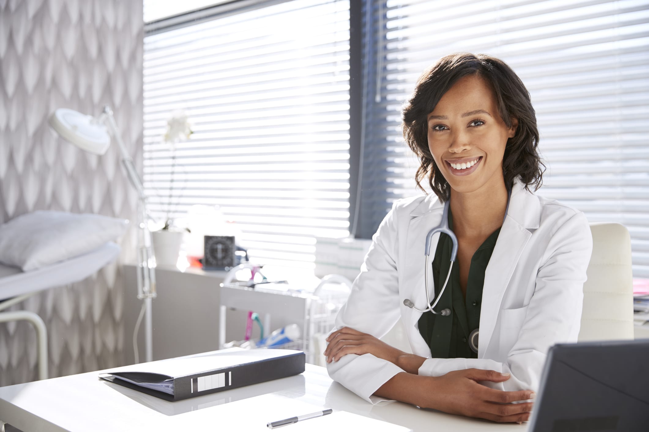Portrait Of Smiling Female Doctor Wearing White Coat With Stethoscope Sitting Behind Desk In Office