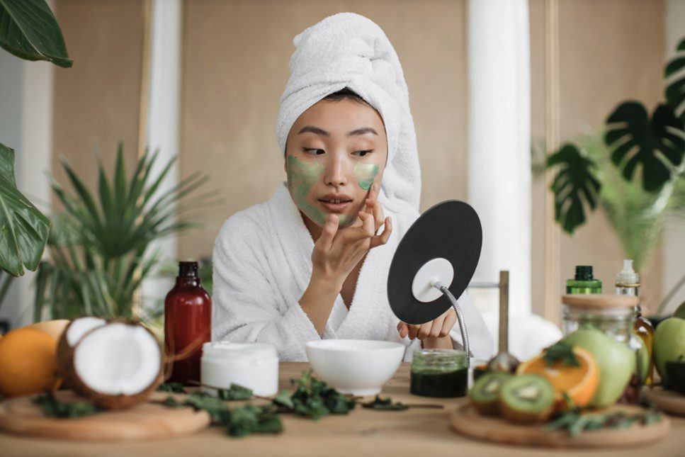 Woman with a facemask on and towel on her head looking in the mirror